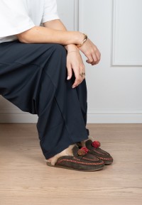 Model wearing brown Poelman NINA slippers with a knitted design, faux fur lining and red pom-poms, paired with navy trousers.