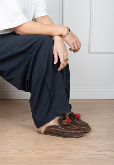 Model wearing brown Poelman NINA slippers with a knitted design, faux fur lining and red pom-poms, paired with navy trousers.