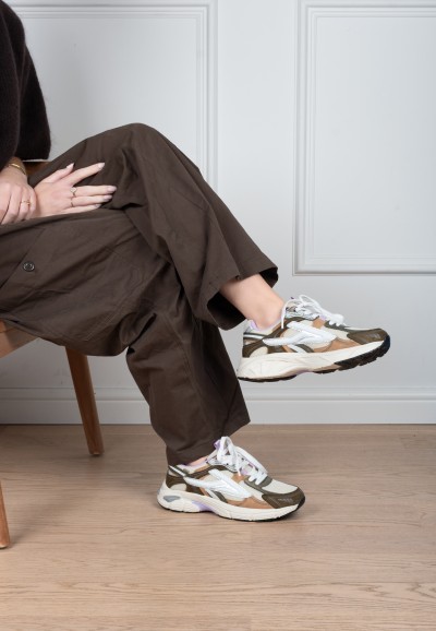 Model draagt POSH By Poelman Ginny wit-goud-lila sneakers met bruine broek – zittend