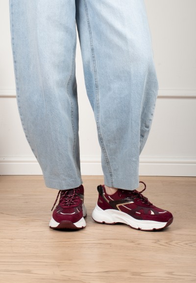 Model trägt POSH By Poelman Ayoki bordeauxfarbene Sneaker mit hellblauer Jeans – Vorderansicht