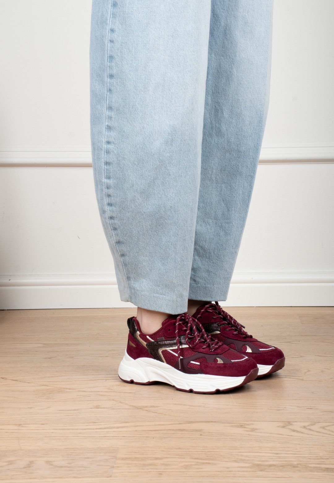 Model trägt POSH By Poelman Ayoki bordeauxfarbene Sneaker mit hellblauer Jeans – Seitenansicht